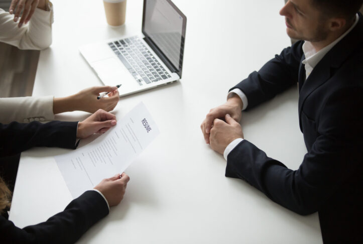 Male applicant having job interview, employers reading resume, asking question. HR representatives hiring young business professional. Recruiting, cooperation, introduction concept. Close up top view